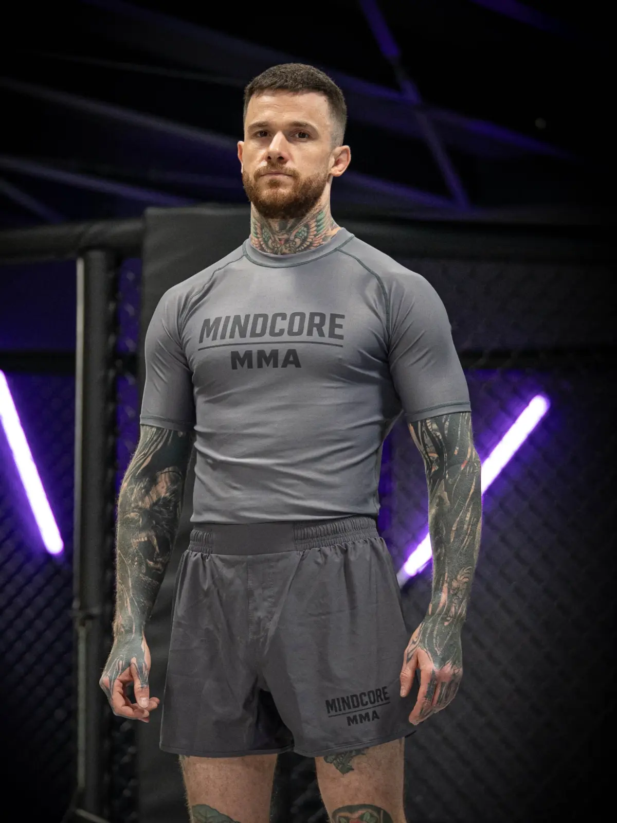 Customer athlete wearing a coordinated grey Mindcore MMA Rash Guard and training shorts beside a cage